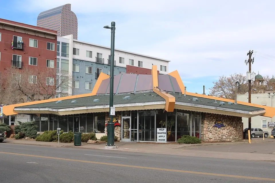The sloped roof of a googie style building is prominently featured in this image of a restaurant in Denver. Tall apartment buildings are seen in the background.